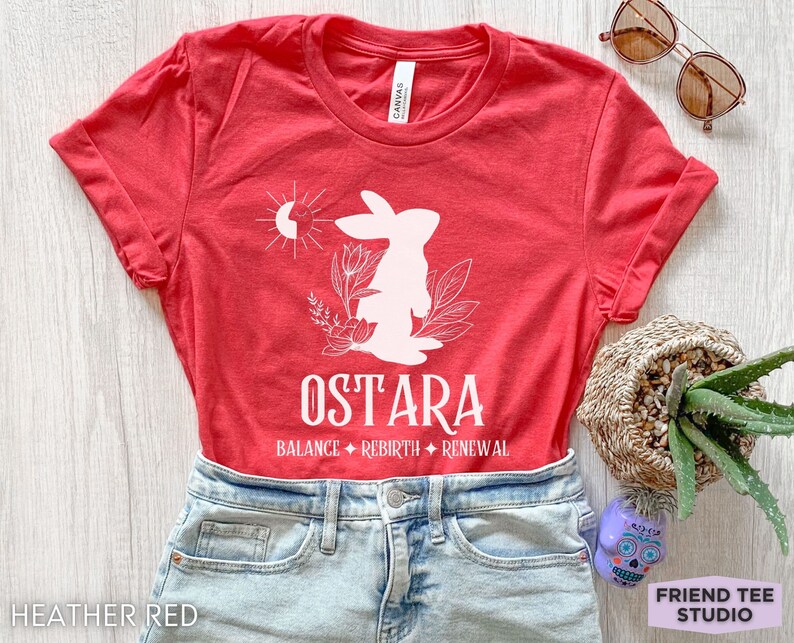 Puede incluir: Una camiseta de color rojo jaspeado con un gr&aacute;fico blanco de un conejo, un sol y elementos florales. El texto dice OSTARA seguido de BALANCE, REBIRTH y RENEWAL. La camiseta se combina con vaqueros azul claro, gafas de sol y una suculenta.