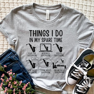 Puede incluir: Una camiseta gris con el texto "THINGS I DO IN MY SPARE TIME" y gráficos relacionados con el saxofón. Los gráficos incluyen un saxofón, un portátil y bocadillos de diálogo. La camiseta se muestra con vaqueros azules y zapatillas negras.
