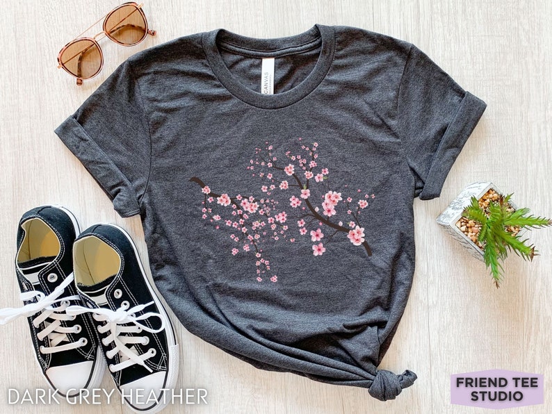 Puede incluir: Una camiseta de color gris oscuro jaspeado con un dise&ntilde;o de rama de flor de cerezo rosa. La camiseta est&aacute; colocada sobre una superficie de madera clara con un par de zapatillas negras y gafas de sol.