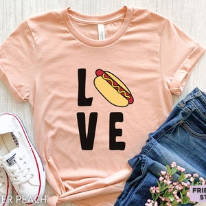 May include: A peach-colored t-shirt with the word "LOVE" in black letters and a cartoon hot dog in the middle of the "O".