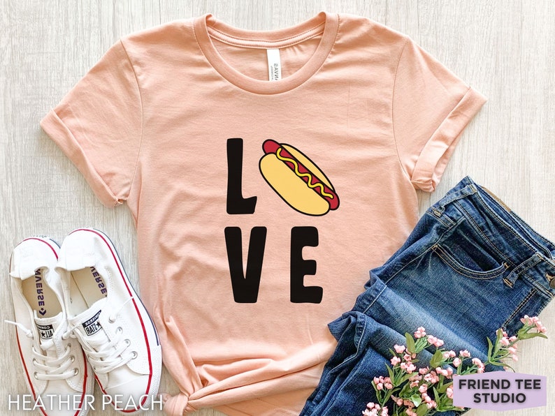 May include: A peach-colored t-shirt with the word "LOVE" in black letters and a cartoon hot dog in the middle of the "O".