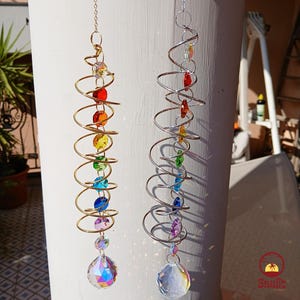 Puede incluir: Dos atrapasoles de cristal colgando de una superficie blanca. Los atrapasoles están hechos de una espiral de metal con cuentas de colores y un prisma de cristal en la parte inferior. Un atrapasol tiene una espiral dorada y el otro una espiral plateada.