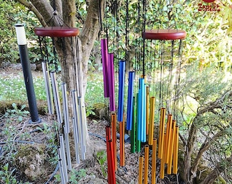 Puede incluir: Un conjunto de carillones de viento coloridos colgando de un árbol. Los carillones tienen tapas de madera y tubos metálicos en plata, morado, azul, verde, naranja y dorado. Están suspendidos por cuerdas negras.