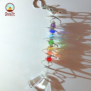 Könnte beinhalten: Ein Regenbogen-Kristall-Sonnenfänger mit einem klaren Kristallprisma, das unten hängt. Der Sonnenfänger besteht aus einer silbernen Drahtspirale mit bunten Perlen.