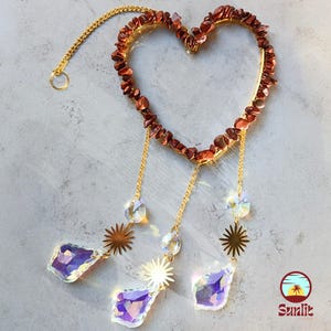 Puede incluir: Un marco de alambre dorado en forma de corazón con cuentas de piedras preciosas rojas, de él cuelgan tres hebras de cadenas dorées con dijes de cristal en forma de sol y gotas de cristal púrpura iridiscente.