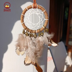 May include: A small brown and white dream catcher with feathers and bells. The dream catcher is hanging from a brown leather cord.