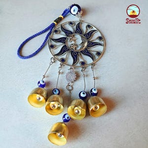 May include: A decorative sun and moon wall hanging with a blue cord and evil eye beads. The metal sun design has blue enamel accents and hangs below five gold-colored bells. The Sunlit logo is in the upper right corner.