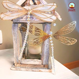 Könnte beinhalten: Zwei Libellen-Windspiele, eines mit schillernden weißen Flügeln und eines mit goldenen Flügeln, hängen an einer Holzlaterne mit einem Glas-Kerzenhalter. Beide Windspiele haben Kristallperlen, die von ihnen herabhängen.