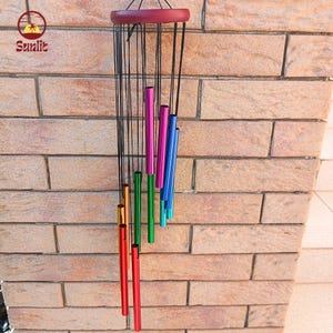 May include: A colorful wind chime with a maroon wooden top and a series of hanging tubes in various colors, including red, orange, green, pink, and blue. The tubes are suspended by black strings. The background is a brick wall.