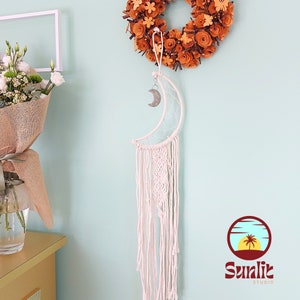 Op de afbeelding: Een witte macramé maan droomvanger met een zilveren halve maan bedel die aan een wit touwtje hangt. De droomvanger hangt aan een lichtblauwe muur met een bruine papieren bloemenkrans erboven.