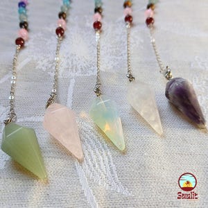 May include: Six crystal pendulums with silver chains hanging from a white surface. The pendulums are made of green, pink, white, clear, and purple crystals.