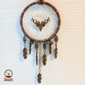 May include: A brown dreamcatcher with a white web and a deer head charm. The dreamcatcher has several bronze feathers and beads hanging from it.