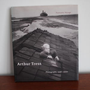 Puede incluir: Una portada de libro de fotografía en blanco y negro titulada "Fantastic Voyage" de Arthur Tress. La portada presenta a una persona joven asomándose desde un agujero en un techo de madera. El título del libro está en texto blanco y el nombre del autor está en texto blanco debajo del título. El texto "Photographs 1956-2000" está en texto blanco en la parte inferior de la portada.