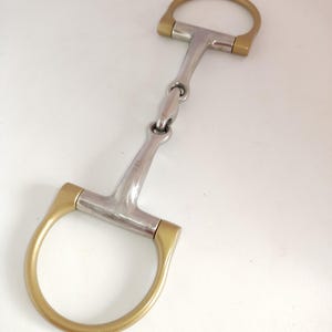 Könnte beinhalten: Ein Pferdegebiss mit einem silberfarbenen Metallmundstück mit Mittelgelenk und zwei goldfarbenen D-förmigen Ringen an den Enden. Die Metalloberfläche ist glatt und reflektierend, mit der Gravur 13,5 cm auf einer Seite des Mundstücks.