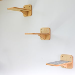 May include: Three wooden cat shelves mounted on a white wall. The shelves are made of light wood and have a simple design. The top shelf is empty, the middle shelf has a small piece of carpet on it, and the bottom shelf is empty.