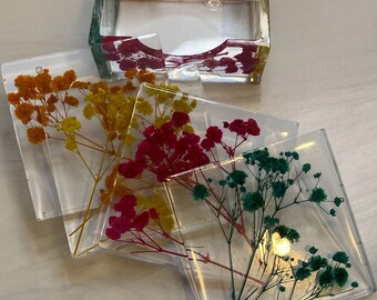 Puede incluir: Cuatro posavasos cuadrados con flores prensadas en diferentes colores. Los posavasos son transparentes y muestran las flores en amarillo, rosa, rojo y verde.