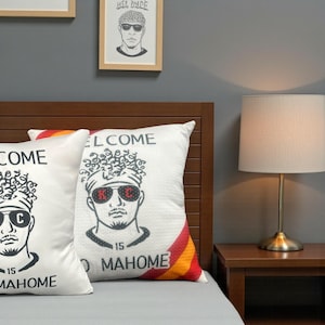 May include: Two white decorative pillows with a graphic of a man wearing sunglasses and the text "WELCOME" and "MAHOME 15". The pillows have a red, orange, and yellow stripe design on the side. A framed drawing hangs above the pillows.
