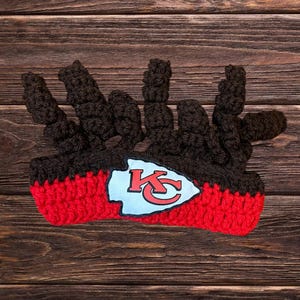 May include: A brown and red crocheted headband with a white and red Kansas City Chiefs logo. The headband is on a brown wooden background.
