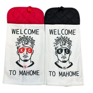 May include: Two white kitchen towels with a black and white illustration of a person wearing sunglasses and a headband. The text "WELCOME TO MAHONE" is printed on the towels. The towels have red and black quilted pot holders attached to the top.