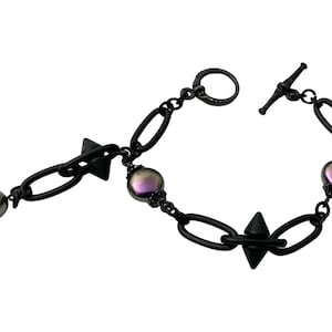 May include: A black bracelet with a toggle clasp, oval chain links, and decorative spikes. The bracelet features iridescent, round beads with purple and green hues. The design is symmetrical and modern.