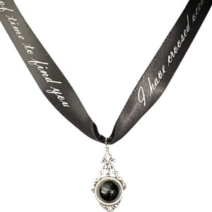 May include: A black ribbon necklace with a silver pendant featuring a black stone. The ribbon has the text "I have crossed oceans of time to find you" printed on it.