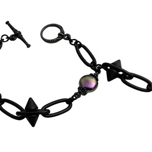 May include: Black metal bracelet featuring oval links, spiked accents, and iridescent pearl-like beads. The bracelet has a toggle clasp and a dangling chain with a single bead. A unique piece of jewelry.