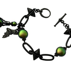 May include: A black bracelet with a toggle clasp, oval links, and cone-shaped spikes. The bracelet features iridescent green orbs and a bat charm. The metal has a matte finish, creating a gothic or alternative aesthetic.
