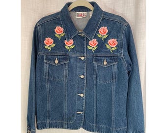 May include: A mid-wash denim jacket featuring embroidered pink roses and green leaves. The jacket has a collared neckline, button-down closure, and two chest pockets. The brand name "Bill Blass" is embroidered on the collar.