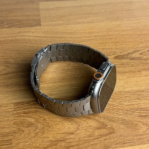 Könnte beinhalten: Eine silberne Apple Watch mit einem schwarzen Armband. Das Armband hat ein einzigartiges, strukturiertes Design.