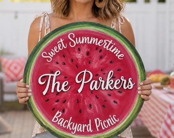Puede incluir: Un letrero redondo de madera con forma de rodaja de sandía con centro rojo, semillas negras y corteza verde. El texto Sweet Summertime, The Parkers y Backyard Picnic aparece en letras blancas sobre el fondo rojo.