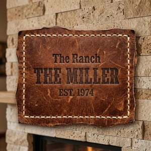 May include: A rectangular, brown leather sign with the text "The Ranch THE MILLER EST. 1974" embossed in dark brown. The sign has a stitched border with white thread and is mounted on a stone wall.
