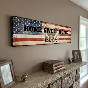 May include: A rectangular wall sign featuring a distressed American flag design with the black text HOME SWEET HOME and EST. 1776. A central eagle emblem is printed over the red and white stripes, and the sign has a weathered finish.