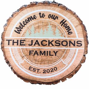 Puede incluir: Un letrero redondo de madera con borde de corteza natural. El letrero dice "Welcome to our Home" y "The Jacksons Family EST. 2020". Un gr&aacute;fico de bosque verde claro est&aacute; en el centro. Un art&iacute;culo de decoraci&oacute;n r&uacute;stica para el hogar.