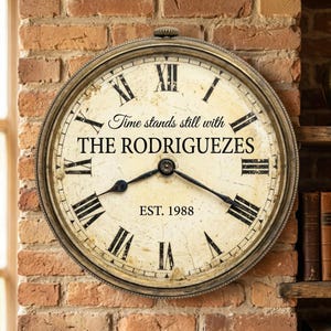 May include: A vintage-style wall clock with a cream-colored face and black Roman numerals. The clock features the text "Time stands still with THE RODRIGUEZES" and "EST. 1988". The clock has a gold-colored metal frame and black clock hands.