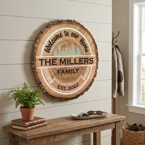 May include: A round wooden sign with a natural bark edge, featuring the text "Welcome to our Home" and "The Millers Family EST. 2022". The sign has a light brown background with a forest graphic and white accents. It is hanging on a light gray wall.