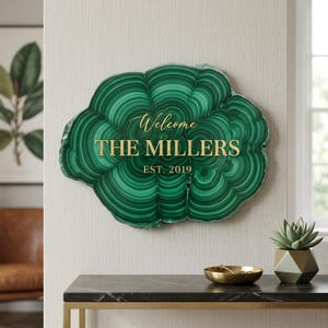May include: A green malachite-style wall hanging with gold lettering that reads "Welcome THE MILLERS EST. 2019". The sign is mounted on a neutral-colored wall. A small gold bowl and a geometric planter are on a nearby table.