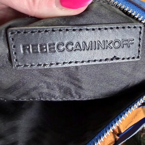 K&ouml;nnte beinhalten: Eine blaue Tasche mit einem schwarzen, rechteckigen Innenaufn&auml;her mit dem gepr&auml;gten Text REBECCAMINKOFF. Die Tasche verf&uuml;gt &uuml;ber einen silberfarbenen Metallrei&szlig;verschluss an der Oberkante und ein schwarzes Stofffutter im Inneren.