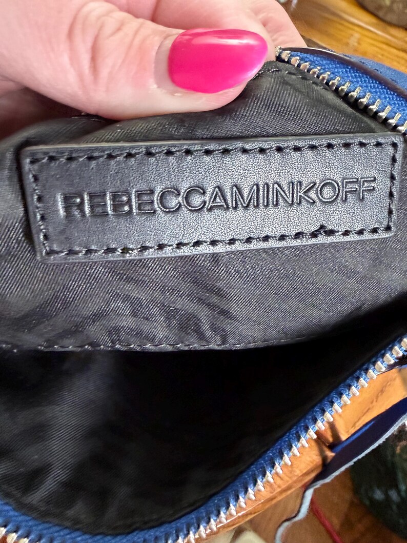 K&ouml;nnte beinhalten: Eine blaue Tasche mit einem schwarzen, rechteckigen Innenaufn&auml;her mit dem gepr&auml;gten Text REBECCAMINKOFF. Die Tasche verf&uuml;gt &uuml;ber einen silberfarbenen Metallrei&szlig;verschluss an der Oberkante und ein schwarzes Stofffutter im Inneren.