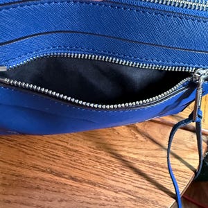 K&ouml;nnte beinhalten: Eine leuchtend blaue Umh&auml;ngetasche mit strukturierter Oberfl&auml;che, ausgestattet mit einem silberfarbenen Metallrei&szlig;verschluss und einem schwarzen Stoffinnenfutter. Die Tasche hat einen passenden blauen Leder-Rei&szlig;verschlussanh&auml;nger.