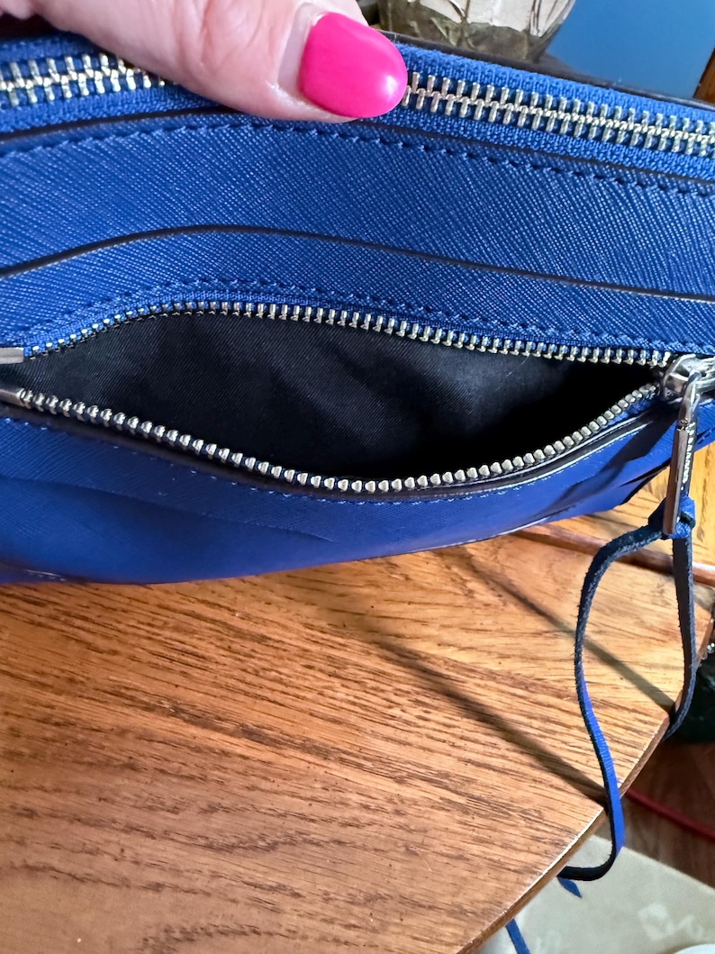 K&ouml;nnte beinhalten: Eine leuchtend blaue Umh&auml;ngetasche mit strukturierter Oberfl&auml;che, ausgestattet mit einem silberfarbenen Metallrei&szlig;verschluss und einem schwarzen Stoffinnenfutter. Die Tasche hat einen passenden blauen Leder-Rei&szlig;verschlussanh&auml;nger.