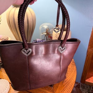 May include: A dark brown leather tote bag featuring two braided leather handles attached to the main body with silver-toned heart-shaped metal hardware. A small silver heart charm hangs from one of the braided handles near the top attachment point.
