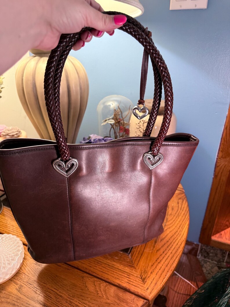May include: A dark brown leather tote bag featuring two braided leather handles attached to the main body with silver-toned heart-shaped metal hardware. A small silver heart charm hangs from one of the braided handles near the top attachment point.
