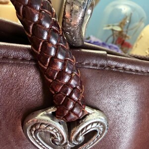 May include: A dark brown leather handbag featuring a braided leather handle attached to a silver-toned metal heart-shaped ornament with intricate engraved detailing. The smooth leather surface of the bag is visible beneath the handle attachment.