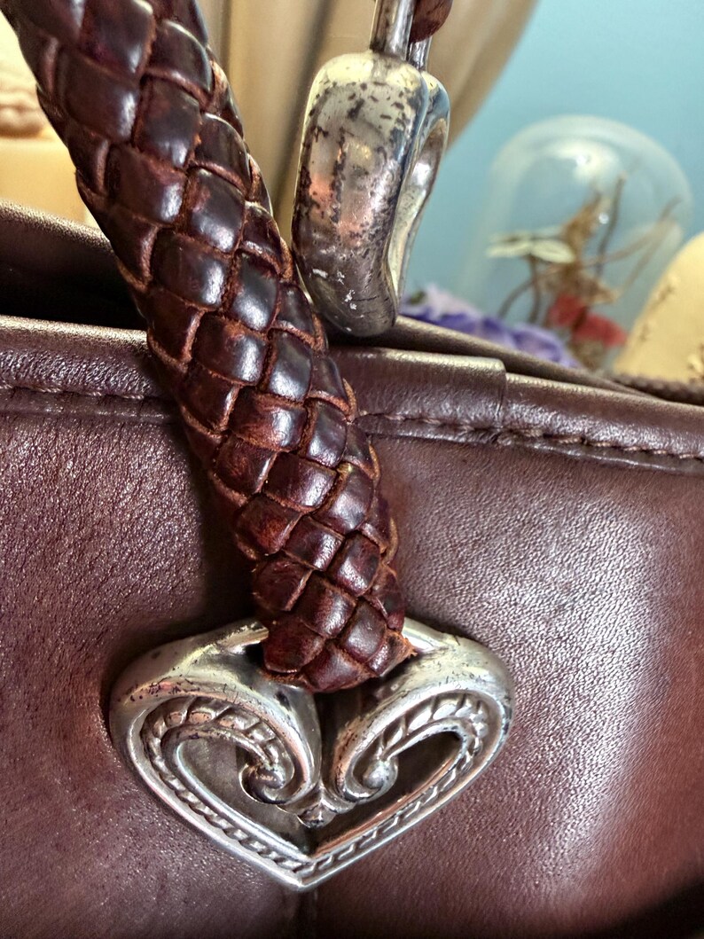 May include: A dark brown leather handbag featuring a braided leather handle attached to a silver-toned metal heart-shaped ornament with intricate engraved detailing. The smooth leather surface of the bag is visible beneath the handle attachment.
