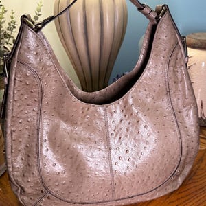 May include: A taupe hobo bag featuring a textured ostrich-embossed finish, a single shoulder strap, and silver-toned metal hardware accents. The bag has a curved silhouette with visible stitching along the front panels and side attachments.