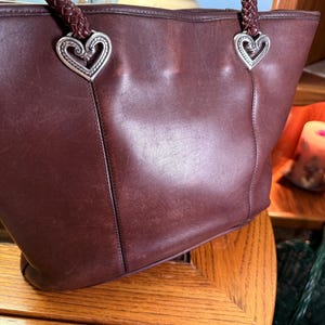 May include: A dark brown leather tote bag featuring two braided leather shoulder straps attached to the bag with silver-toned metal heart-shaped hardware. The bag has a smooth, matte finish with visible stitching details along the front panel.