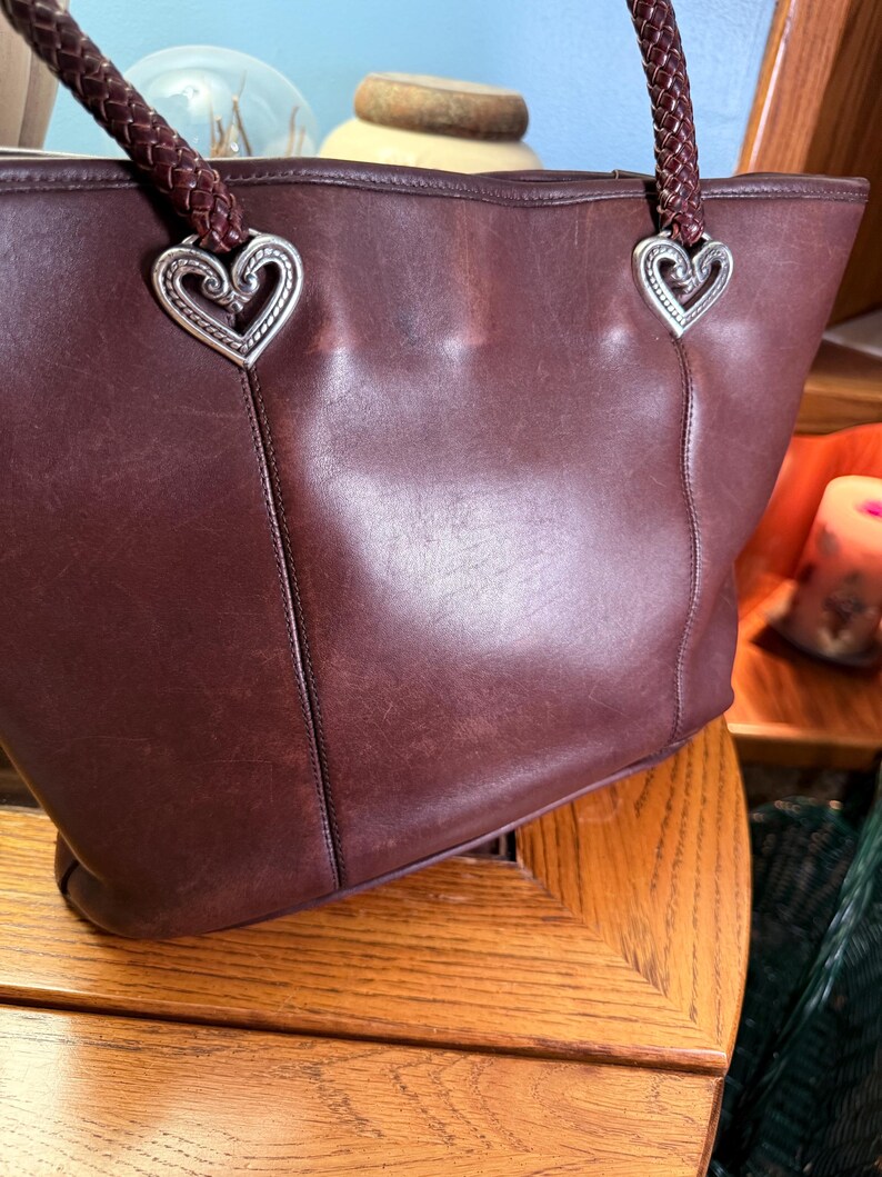 May include: A dark brown leather tote bag featuring two braided leather shoulder straps attached to the bag with silver-toned metal heart-shaped hardware. The bag has a smooth, matte finish with visible stitching details along the front panel.