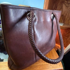May include: A dark brown leather tote bag featuring two braided leather handles attached to the body with silver-toned heart-shaped metal hardware. The bag has a smooth, matte finish and a structured rectangular shape with visible stitching.