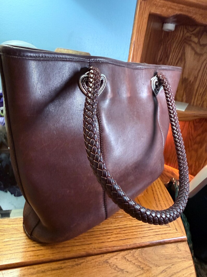 May include: A dark brown leather tote bag featuring two braided leather handles attached to the body with silver-toned heart-shaped metal hardware. The bag has a smooth, matte finish and a structured rectangular shape with visible stitching.