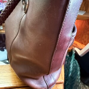 May include: A dark brown leather tote bag featuring braided leather handles attached with silver-toned metal hardware. The bag has visible vertical stitching along the side panel and a smooth, matte finish on the exterior leather surface.